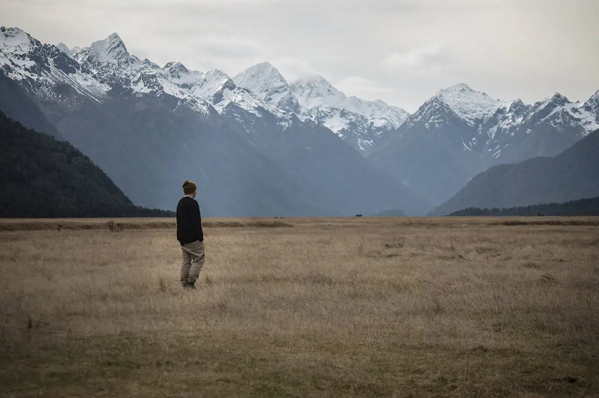 Eglinton Valley: ULTIMATE Guide For Visiting (2026) 21 A man gazes into the distance while standing in a wide expanse of dry grass, with towering snow-capped mountains creating a breathtaking backdrop. The tranquil setting of Eglinton Valley emphasizes the natural beauty of the area.