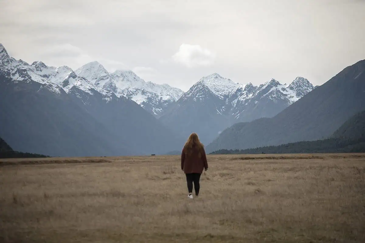 Eglinton Valley: ULTIMATE Guide For Visiting (2026) 22 tasha amy with long hair walks away from the camera across an expansive field, with the dramatic, snow-capped peaks of Eglinton Valley rising in the distance under a cloudy sky.