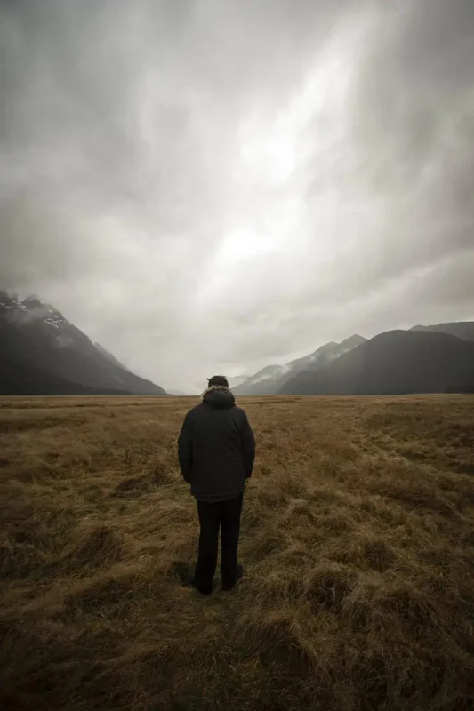 Eglinton Valley: ULTIMATE Guide For Visiting (2026) 26 A person in a dark coat stands facing away, looking towards the expansive plains and misty mountains in the distance. The overcast sky and muted tones create a reflective and tranquil mood in Eglinton Valley.