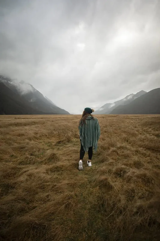 Eglinton Valley: ULTIMATE Guide For Visiting (2026) 17 tasha amy dressed in a green poncho and white sneakers walks across the dry grass, surrounded by towering mountains and an overcast sky. The misty peaks in the distance add to the dramatic scenery of Eglinton Valley.