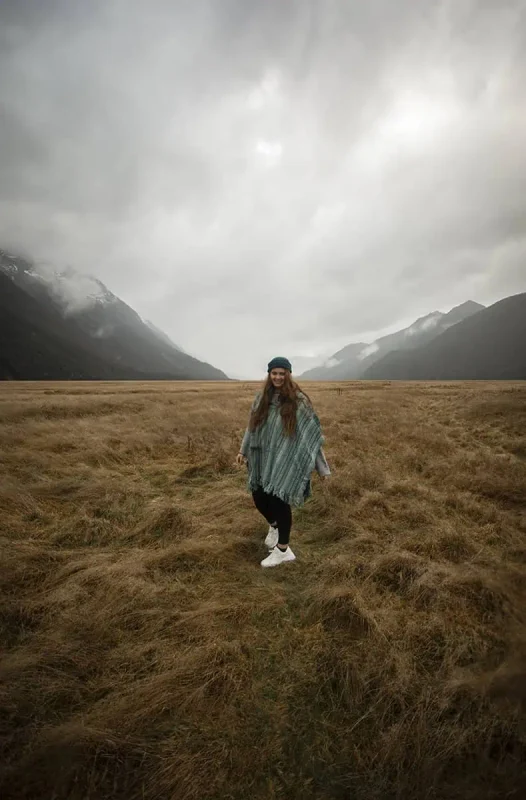 Eglinton Valley: ULTIMATE Guide For Visiting (2026) 16 tasha amy in a green poncho smiles while standing on a vast, grassy plain, with mist-covered mountains in the background. The scene captures the serene and breathtaking landscape of Eglinton Valley.