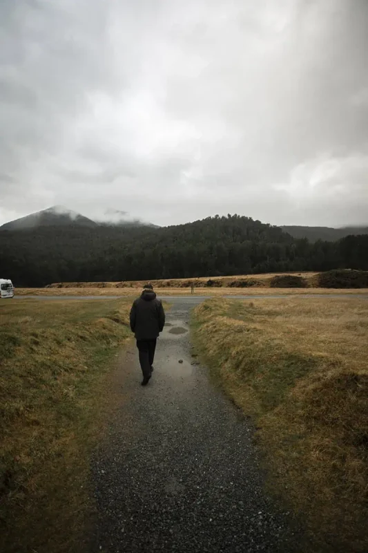 Eglinton Valley: ULTIMATE Guide For Visiting (2026) 24 A person in a dark coat walks along a gravel path leading into the distance, bordered by grassy fields and distant forested hills under a cloudy sky. The peaceful solitude of the scene is accentuated by the misty weather.