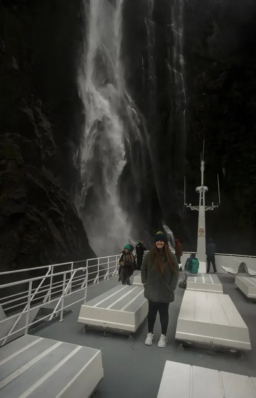 Mirror Lakes: Is It Worth Visiting In Milford Sound, NZ? 25 A group of people on a boat deck near a powerful waterfall in Milford Sound. The waterfall cascades down a steep, rocky cliff, creating a misty spray that envelops the area. The passengers, dressed warmly, seem captivated by the natural spectacle.