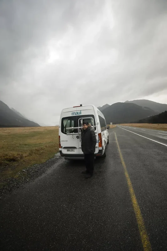 Mirror Lakes: Is It Worth Visiting In Milford Sound, NZ? 23 A person standing next to a white "Maui" campervan on a wet, overcast day. The campervan is parked on a roadside with a vast, open field and misty mountains in the background. The gray sky and damp ground create a moody, traveler's scene.
