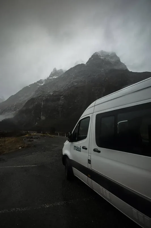 Mirror Lakes: Is It Worth Visiting In Milford Sound, NZ? 24 A white "Maui" campervan parked near towering, mist-covered mountains. The rugged peaks rise dramatically against the overcast sky, creating a sense of isolation and adventure. The wet road and surrounding landscape emphasize the wild, untamed beauty of Milford Sound.