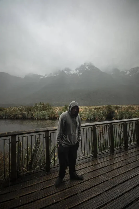 Mirror Lakes: Is It Worth Visiting In Milford Sound, NZ? 19 A person in a gray hoodie stands on a wooden platform overlooking Mirror Lakes in Milford Sound. The platform is surrounded by dense greenery and offers a view of the misty, snow-capped mountains in the background. The gray, cloudy sky and the reflective surface of the lake create a serene and contemplative atmosphere.