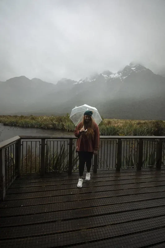 Mirror Lakes: Is It Worth Visiting In Milford Sound, NZ? 20 tasha amy with long hair and a red jacket, holding a transparent umbrella, stands on a wooden platform by a lake. The backdrop features a stunning view of mist-covered mountains and dense vegetation. The overcast sky and light drizzle add a touch of melancholy to the otherwise peaceful setting.