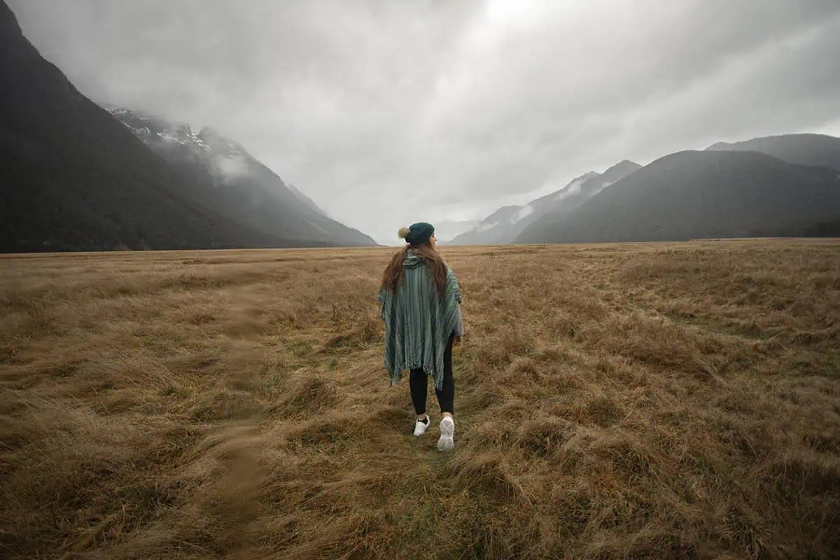 Mirror Lakes: Is It Worth Visiting In Milford Sound, NZ? 27 tasha amy wearing a green poncho and a beanie walks across a vast, golden field towards mist-shrouded mountains in the distance. The wide-open space and towering mountain ranges create a sense of grandeur and adventure, while the overcast sky adds an element of solitude and introspection.