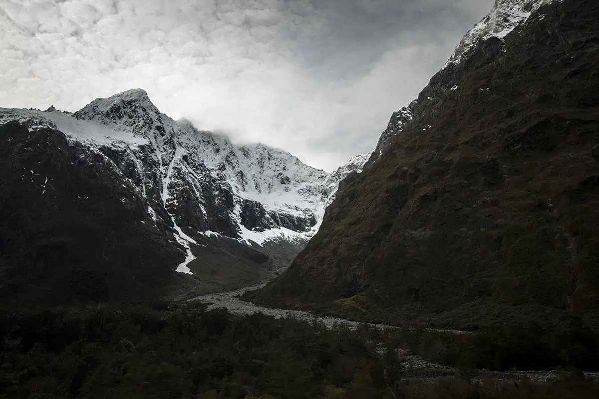 Mirror Lakes: Is It Worth Visiting In Milford Sound, NZ? 28 A narrow valley flanked by steep, snow-covered mountains with a rocky riverbed cutting through the middle. The rugged landscape is framed by towering cliffs and a cloudy sky, emphasizing the dramatic and untamed beauty of the natural surroundings. The stark contrast between the snow and the rocky terrain highlights the harshness and magnificence of the environment.