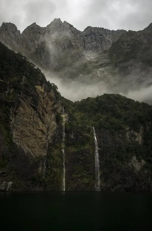 Mirror Lakes: Is It Worth Visiting In Milford Sound, NZ? 26 Tall, rugged cliffs with cascading waterfalls are partially shrouded in mist, creating a dramatic and ethereal scene. The lush green foliage contrasts with the dark, rocky surfaces, while the waterfalls add a sense of movement and tranquility to the landscape.