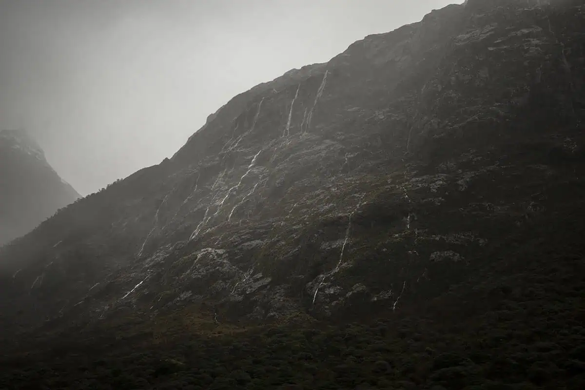 Monkey Creek: BEST Stop On The Way To Milford Sound 19 A close-up shot of the rocky mountainside with waterfalls forming on a rainy day. The steep slope is streaked with white, indicating recent snow or water runoff. The overcast sky and misty atmosphere create a dramatic and moody scene, emphasizing the rugged beauty of the landscape.