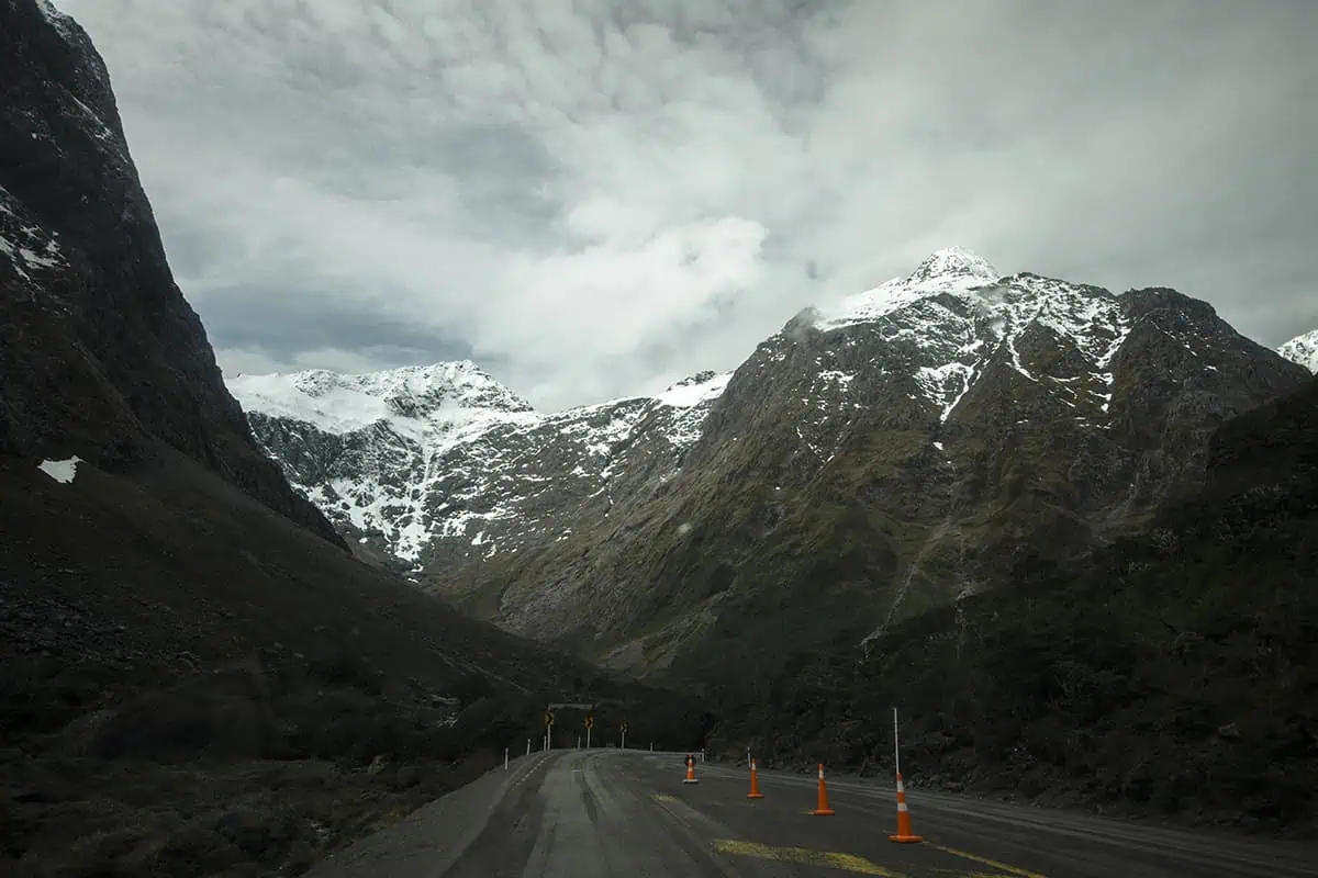 Monkey Creek: BEST Stop On The Way To Milford Sound 20 A scenic road through to Milford Sound in New Zealand, winds through a mountainous landscape with towering, snow-capped peaks. Bright orange traffic cones line the roadside, adding a splash of color to the natural setting. The mountains' rugged surfaces are partially covered in snow, and the sky is filled with thick, low-lying clouds, creating a dramatic backdrop.