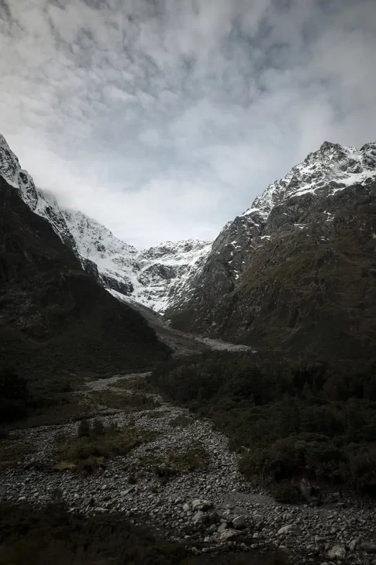 Monkey Creek: BEST Stop On The Way To Milford Sound 33 A rocky streambed winds its way down the mountain at Monkey Creek. The stream is flanked by dense vegetation and trees, leading up to the steep, snow-dusted slopes of the surrounding mountains. The cloudy sky above adds to the dramatic and remote feel of the area, highlighting the untouched wilderness.
