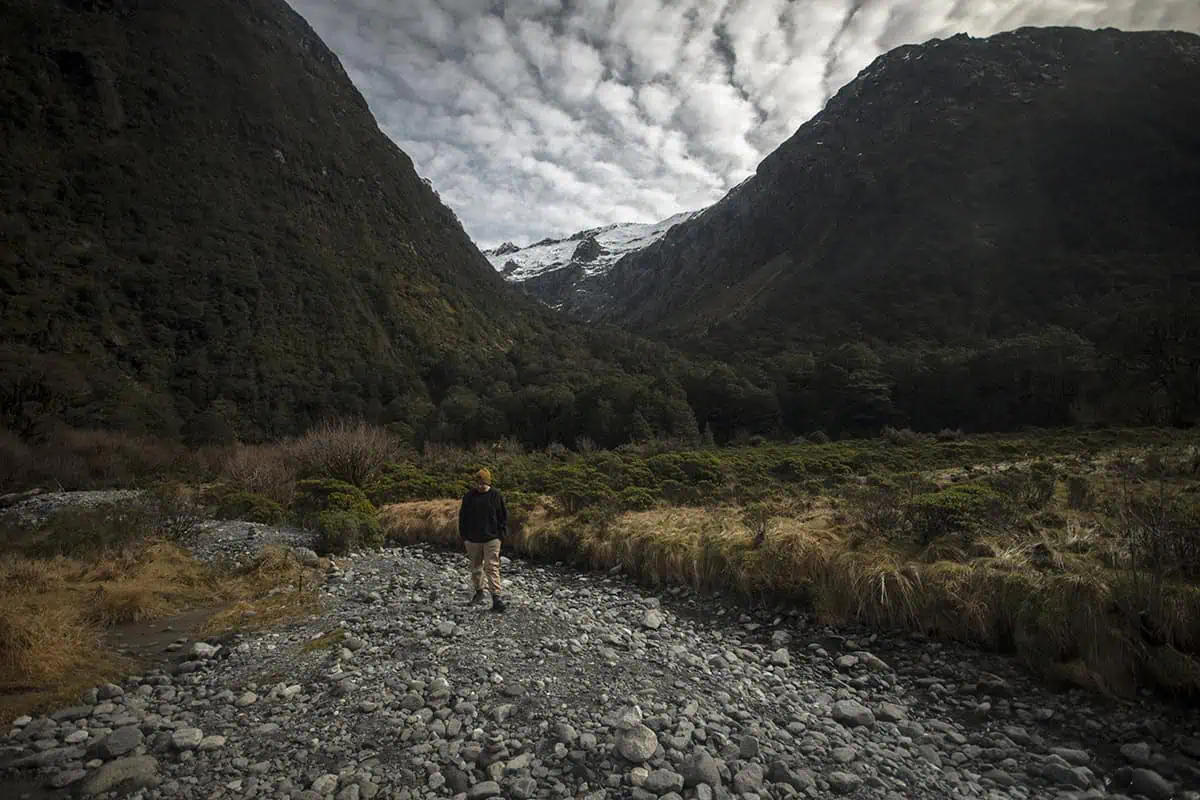 Monkey Creek: BEST Stop On The Way To Milford Sound 26 A lone hiker walks along a rocky path at Monkey Creek, New Zealand, surrounded by a dense forest and steep, snow-capped mountains. The person, wearing a dark jacket and yellow beanie, appears small against the vast, dramatic landscape. The sky above is filled with clouds, adding to the sense of solitude and wilderness.