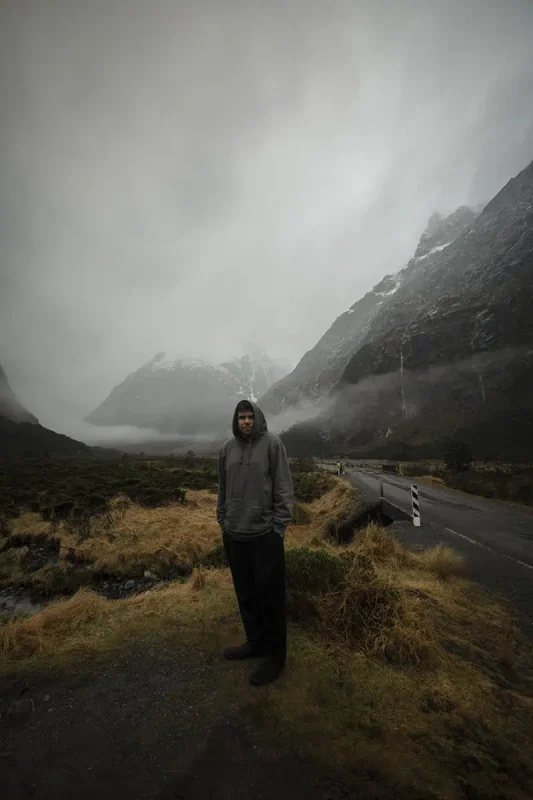 Monkey Creek: BEST Stop On The Way To Milford Sound 18 A man in a grey hoodie stands on a grassy, rocky path at Monkey Creek, New Zealand. Behind him, a winding road disappears into the misty, mountainous background. The dark, imposing mountains are partially obscured by low-hanging clouds, creating a mysterious and atmospheric scene.