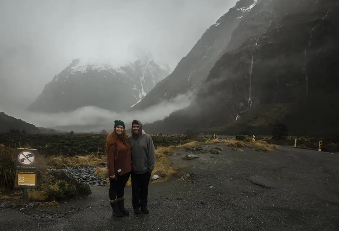 Monkey Creek: BEST Stop On The Way To Milford Sound 16 A couple poses together at Monkey Creek, New Zealand, with the woman wearing a rust-colored sweater and beanie and the man in a grey hoodie. They stand in front of a dramatic landscape of mist-covered mountains and a lush valley. A sign in the foreground advises against feeding the local kea birds, adding a touch of local wildlife awareness to the scene.