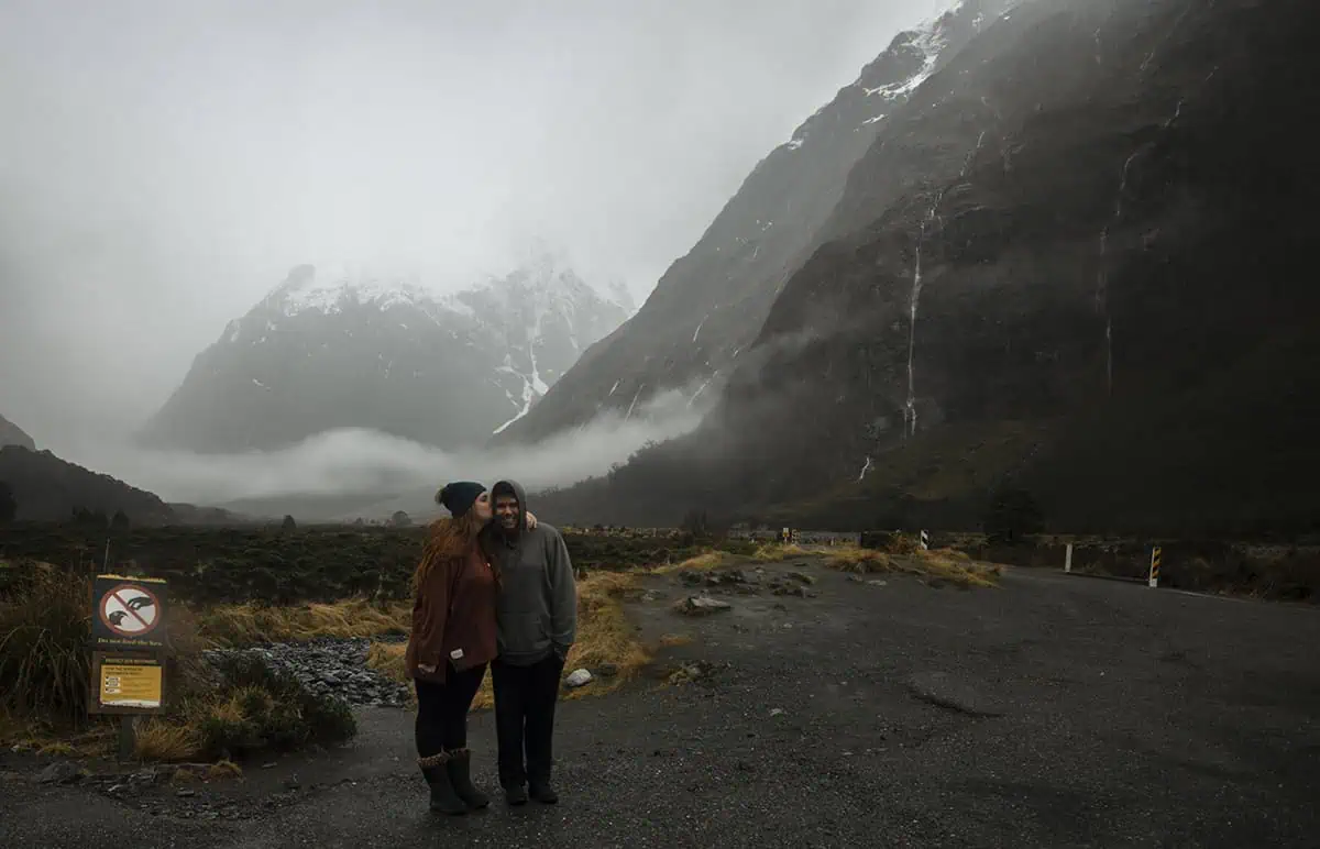 Monkey Creek: BEST Stop On The Way To Milford Sound 12 tasha amy and her partner james stand close together at Monkey Creek New Zealand. The woman kisses the man's cheek as they smile in front of the dramatic, mist-covered mountains. The overcast sky and rugged landscape provide a stunning natural backdrop, emphasizing the area's remote beauty