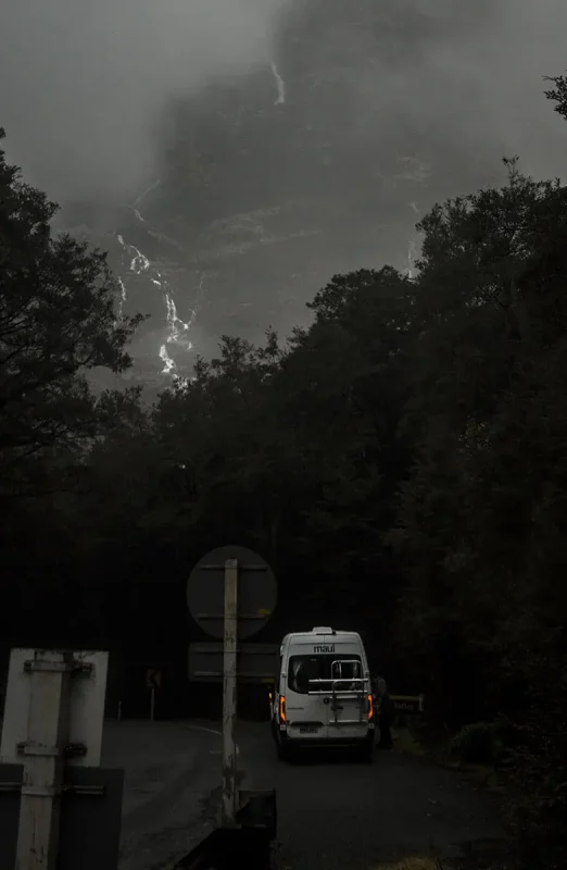 A white camper van marked "maui" drives on a misty, forested road, with the faint outline of waterfalls visible on the mountain slope ahead. The scene evokes a sense of adventure and exploration in Fiordland National Park.