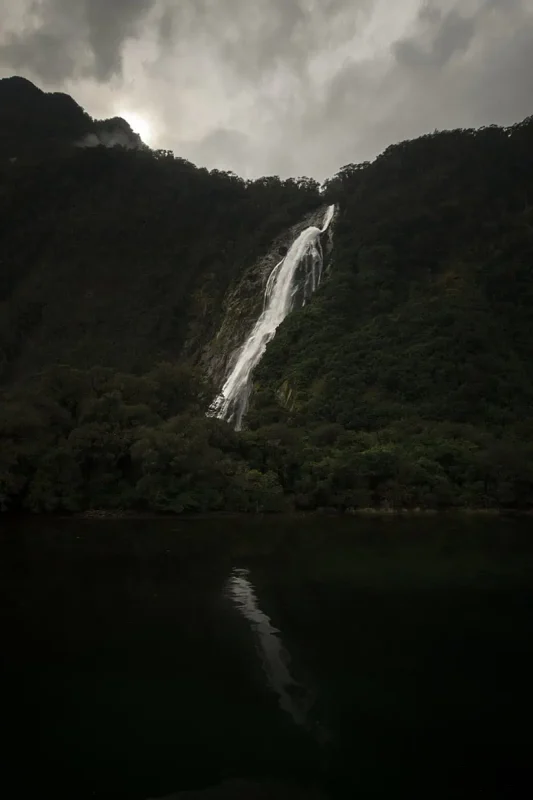 Milford Sound In The Rain: The BEST Time To Visit 2026 17 A powerful bowen waterfall streams down a steep, forested mountain slope, surrounded by dense greenery. The sky is overcast, adding a moody atmosphere to the scene, typical of Milford Sound’s frequent rains.