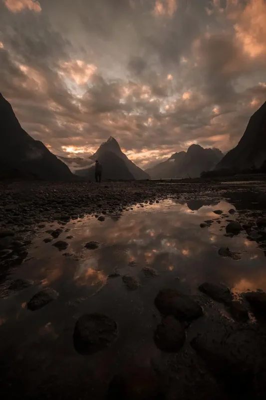Milford Sound In The Rain: The BEST Time To Visit 2026 38 reflective pool with Milford Sound's iconic Mitre Peak in the background at sunset. The sky is ablaze with orange and pink hues, reflected in the still waters, creating a serene yet dramatic scene.