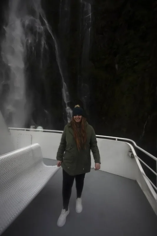 Milford Sound In The Rain: The BEST Time To Visit 2026 24 tasha amy in a green parka stands on a boat deck near a powerful waterfall in Milford Sound. The waterfall creates a misty spray, and the overcast sky adds to the sense of adventure and the typical wet weather of the region.
