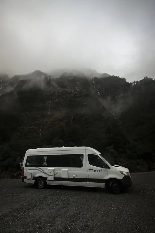 Milford Sound In The Rain: The BEST Time To Visit 2026 28 A white camper van is parked against the backdrop of a misty, mountainous landscape in Milford Sound. The mountain is partially obscured by low clouds, with thin waterfalls trickling down its slopes, creating a picturesque yet typical rainy day in Milford Sound.