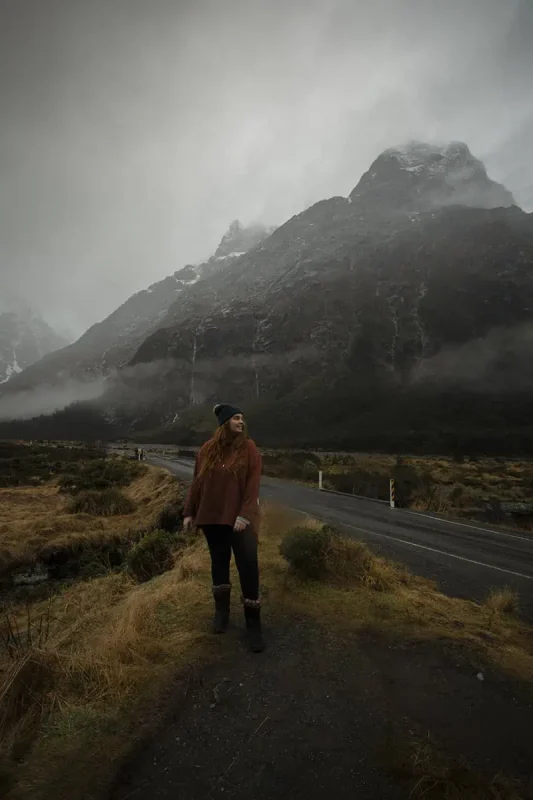 Milford Sound In The Rain: The BEST Time To Visit 2026 32 tasha amy stands by a roadside in Milford Sound, with mist-shrouded mountains in the background. The rugged terrain and the overcast sky create a dramatic and atmospheric scene, typical of the rain in Milford Sound.