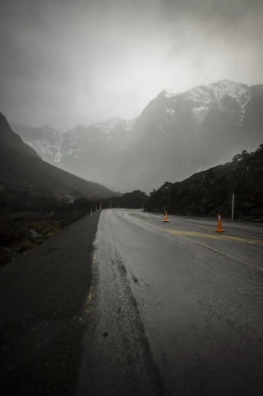 Milford Sound In The Rain: The BEST Time To Visit 2026 30 A wet road winds through a misty mountain landscape in Milford Sound. The road is lined with orange traffic cones, and the overcast sky and low-hanging clouds create a moody and serene atmosphere.