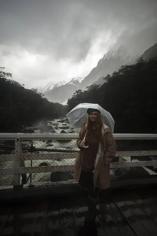 Milford Sound In The Rain: The BEST Time To Visit 2026 22 tasha amy stands on a bridge holding an umbrella, overlooking a fast-flowing river surrounded by misty mountains in Milford Sound. The overcast sky and low clouds highlight the typical rainy weather in the region.