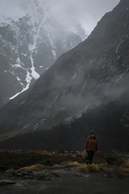 Milford Sound In The Rain: The BEST Time To Visit 2026 18 tasha amy in a brown jacket stands against a backdrop of towering, misty mountains in Milford Sound. The scene is dominated by grey skies and a light drizzle, capturing the essence of exploring the region during the rain.