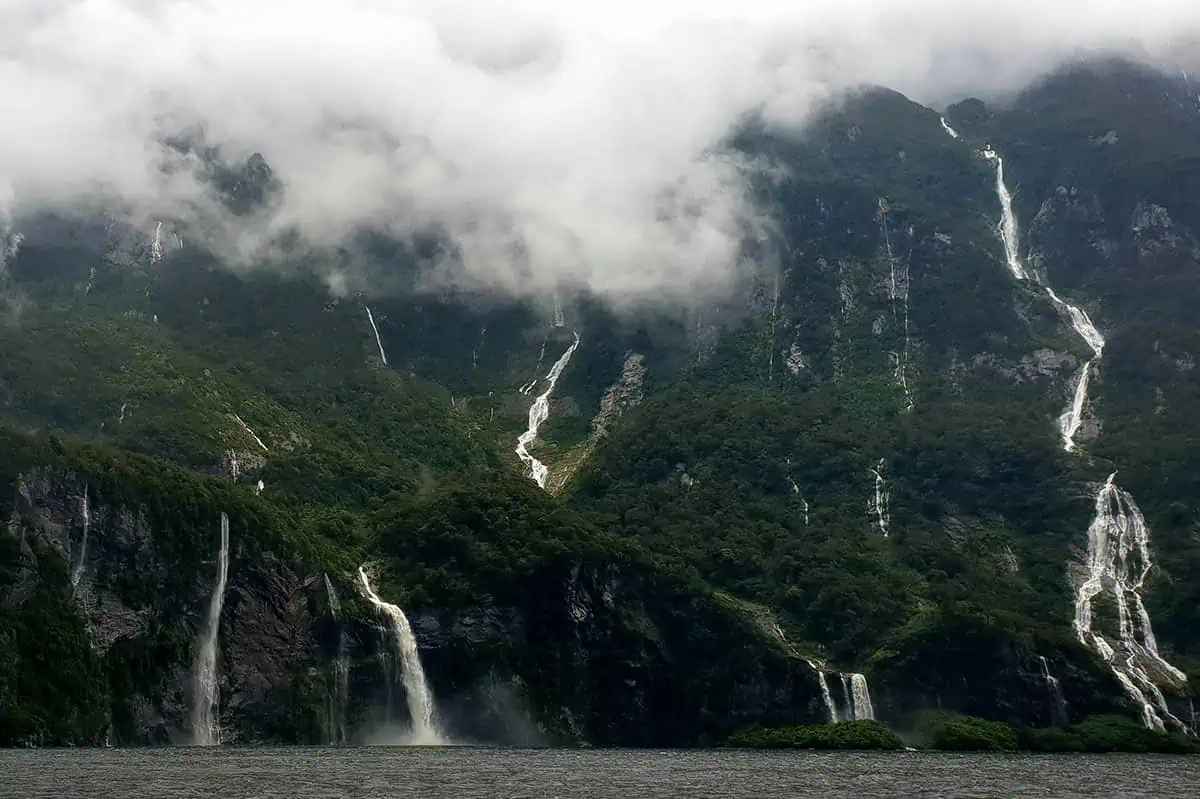 Milford Sound In The Rain: The BEST Time To Visit 2026 1 aterfalls cascade down lush, green mountains shrouded in mist and low-hanging clouds in Milford Sound. The dramatic scenery captures the raw beauty of the landscape, with multiple waterfalls visible against the dense foliage. This image perfectly showcases the ethereal atmosphere of rain in Milford Sound.