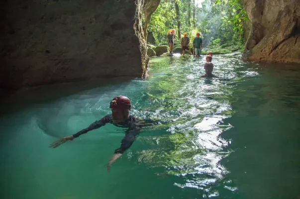 A group of people, equipped with helmets and headlamps, explore an emerald green water-filled cave. In the foreground, a person swims towards the camera, stretching their arms forward. The background reveals a small group standing on rocks at the cave's mouth, bathed in natural light filtering through the lush jungle foliage. This scene captures the thrill and beauty of solo travel in Belize, showcasing the adventure of exploring natural cave systems.