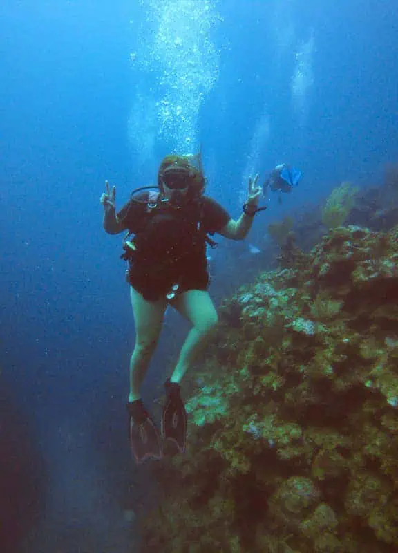 tasha amy floats near a coral reef, giving a peace sign with both hands while surrounded by clear blue water. The diver’s attire includes a black wetsuit, fins, and a diving mask, with bubbles rising towards the surface. Colorful coral formations and a variety of fish are visible in the background. This underwater snapshot captures the adventurous spirit and the vibrant marine life explored in Belize.