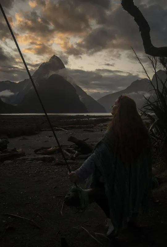 Milford Sound In Winter: Is It Still Worth The Hype? 33 tasha amy sits on a rustic swing made of a wooden log and ropes, set against the backdrop of Milford Sound at dusk. The sky is painted with vibrant shades of orange and purple, reflecting on the calm water. The mist-covered peaks add to the dramatic and serene atmosphere of winter in Milford Sound.