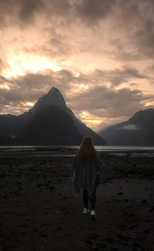 Milford Sound In Winter: Is It Still Worth The Hype? 34 tasha amy walks across a pebbly shore, their back turned to the camera. The sky is illuminated by a warm, golden sunset, casting a soft glow over the misty mountains and calm waters of Milford Sound. The scene captures the tranquility and beauty of the fjord during winter.