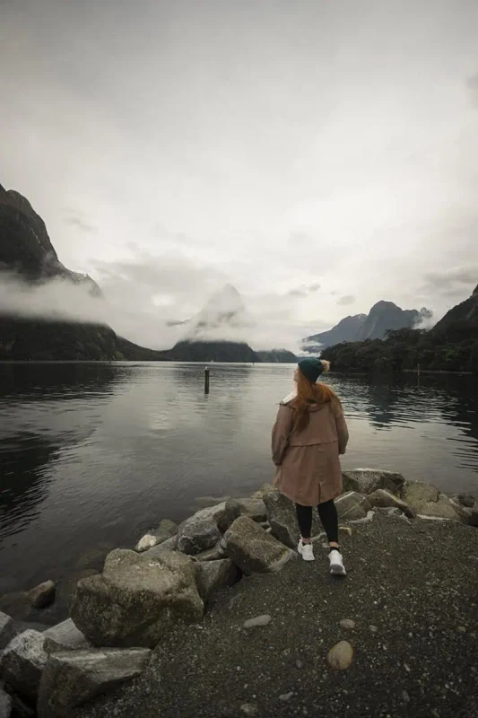 Milford Sound In Winter: Is It Still Worth The Hype? 27 tasha amy stands on a rocky outcrop by the water's edge, wearing a tan coat and dark beanie. The calm waters of Milford Sound reflect the mist-covered peaks in the distance, creating a serene and contemplative winter scene in Milford Sound.