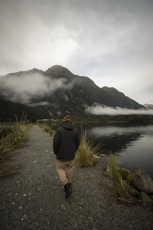 Milford Sound In Winter: Is It Still Worth The Hype? 23 A person in a dark coat and mustard-yellow beanie walks along a gravel path beside a calm, misty lake. The towering, cloud-covered mountains in the background create a dramatic and serene atmosphere, typical of winter in Milford Sound.