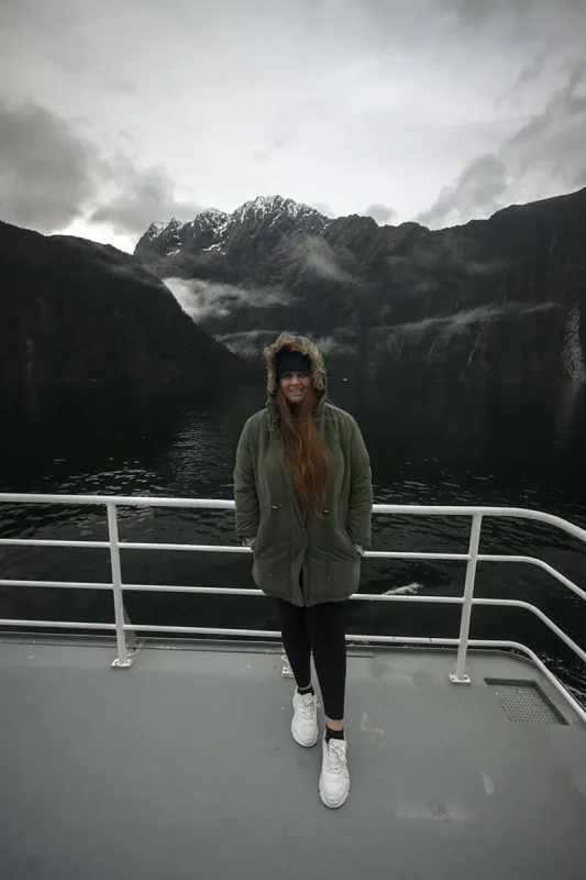 Milford Sound In Winter: Is It Still Worth The Hype? 17 tasha amy stands on the deck of a boat, smiling at the camera, wrapped in a green parka with a fur-lined hood. The dark, tranquil waters of Milford Sound surround the boat, and towering, snow-capped mountains rise majestically in the background. The overcast sky and mist add to the serene and cool ambiance of winter in Milford Sound.