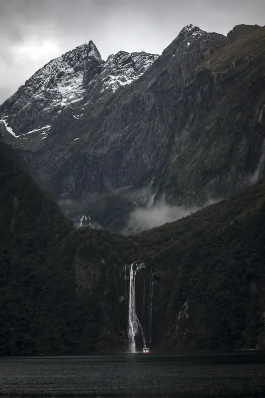 Milford Sound In Winter: Is It Still Worth The Hype? 18 A distant view of a cascading waterfall set against the dramatic backdrop of Milford Sound's towering cliffs and rugged, snow-capped peaks. A small boat is visible at the base of the waterfall, highlighting the immense scale and natural beauty of the winter landscape.