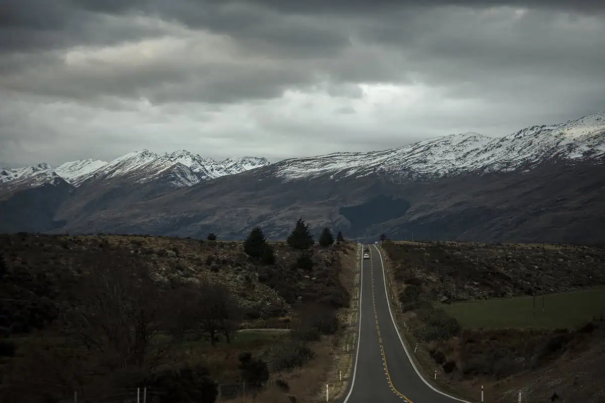 Milford Sound In Winter: Is It Still Worth The Hype? 32 A long, straight road stretches into the distance, flanked by rugged, snow-dusted hills and mountains. The cloudy sky and barren landscape create a stark, dramatic scene, characteristic of a winter drive through the Milford Sound region.