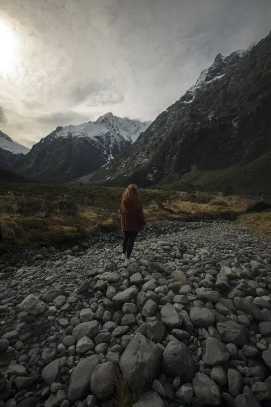 Milford Sound In Winter: Is It Still Worth The Hype? 24 tasha amy walks alone on a bed of rocks in a valley surrounded by imposing, snow-covered mountains. The expansive, serene landscape and overcast sky capture the rugged and awe-inspiring beauty of winter in Milford Sound.