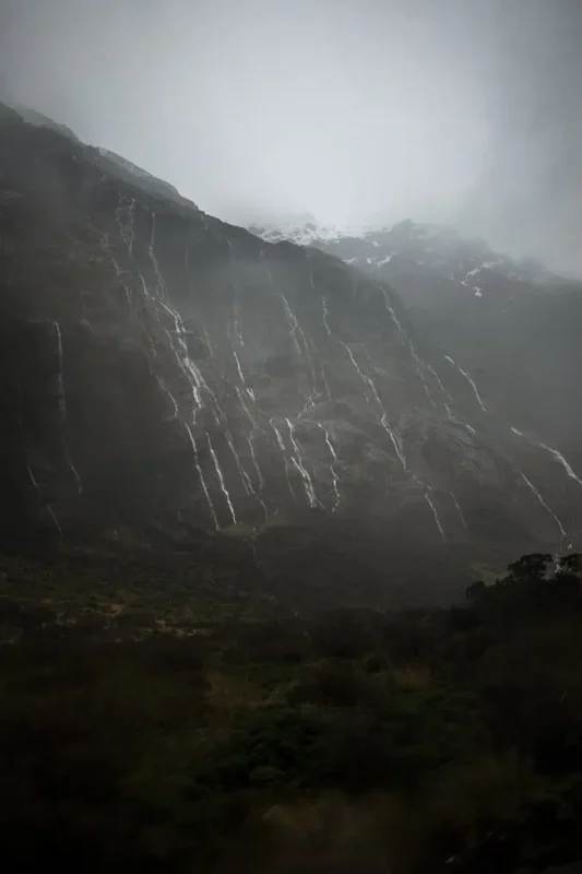 Milford Sound In Winter: Is It Still Worth The Hype? 20 Steep, rugged mountains draped in mist and rain, with multiple thin waterfalls cascading down their slopes. The scene is dramatic and moody, highlighting the raw natural beauty and frequent rains typical of Milford Sound's winter season.