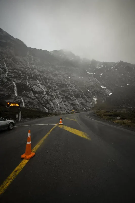 Milford Sound In Winter: Is It Still Worth The Hype? 21 A winding mountain road with orange traffic cones and a road sign. The rocky cliffs on either side are covered with streaks of rainwater and patches of snow. The overcast sky and fog add to the chilly, rugged atmosphere of the winter season in Milford Sound.