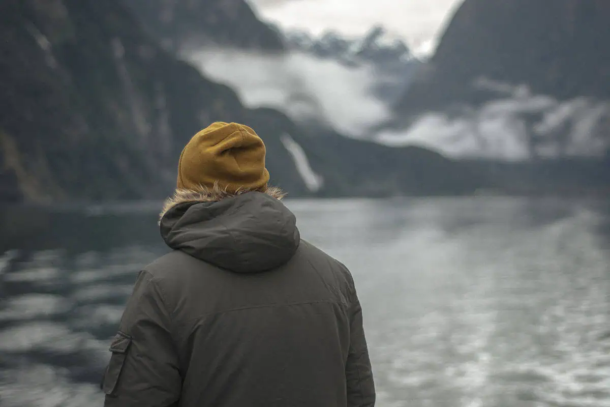 Milford Sound In Winter: Is It Still Worth The Hype? 9 A person with their back to the camera stands on the edge of a body of water, wearing a mustard-yellow beanie and a dark grey parka with a fur-lined hood. The overcast sky and mist shroud the towering mountains in the background, creating a serene and contemplative atmosphere typical of winter in Milford Sound.