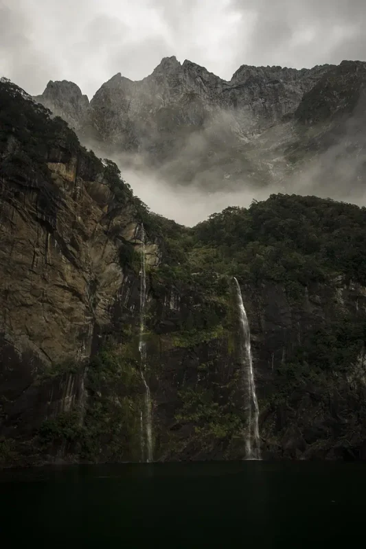 Fiordland & Milford Sound Lord Of The Rings Locations (2026) 17 Twin waterfalls cascade down steep cliffs into dark water with mist clinging to the rocky mountains in Milford Sound, New Zealand
