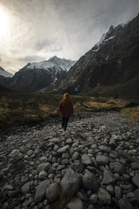 Fiordland & Milford Sound Lord Of The Rings Locations (2026) 18 tasha amy walks across a rocky riverbed surrounded by towering snow-capped mountains in Fiordland, New Zealand