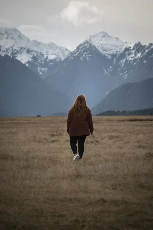 Fiordland & Milford Sound Lord Of The Rings Locations (2026) 32 tasha amy with long hair in a red jacket walks across an open golden field toward sharp snowy peaks in eglinton valley