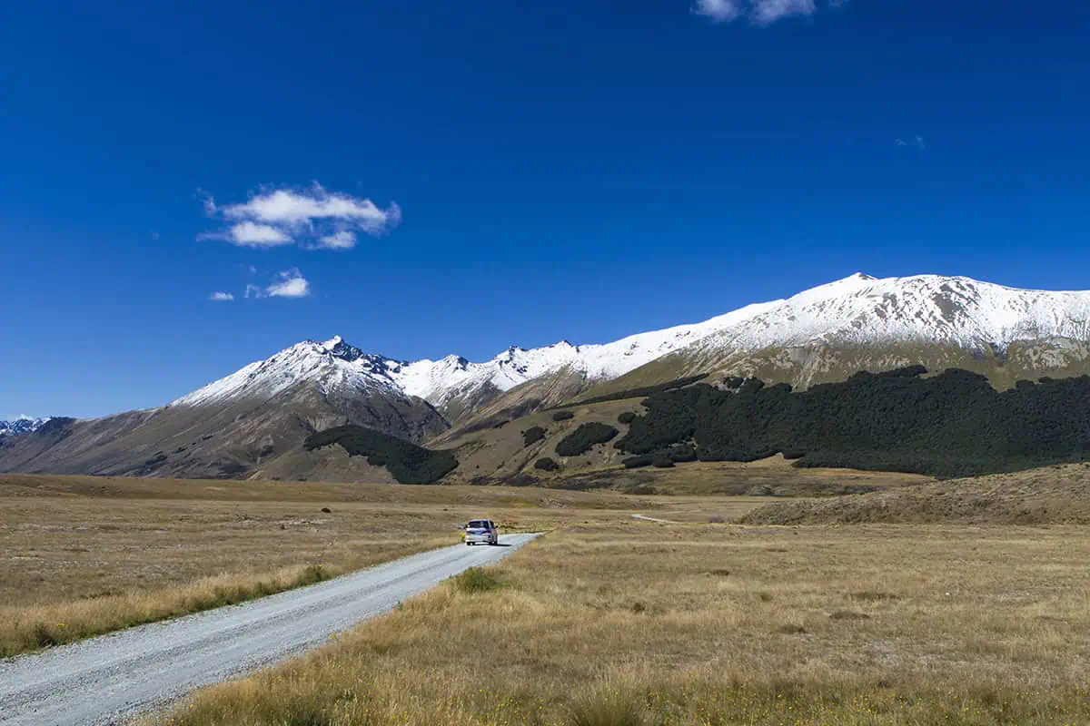 Fiordland & Milford Sound Lord Of The Rings Locations (2026) 19 A car drives along a gravel road toward snow-capped peaks under a blue sky in New Zealand’s Lord of the Rings landscape.