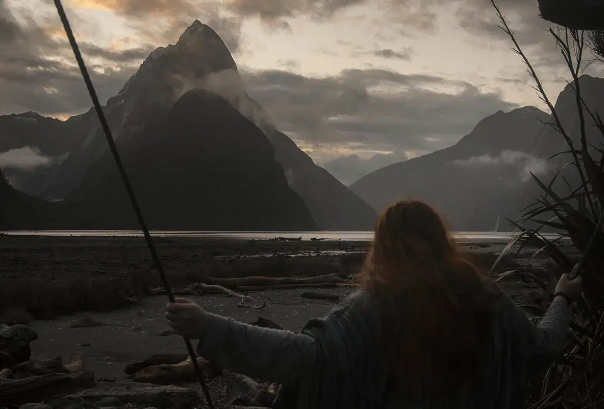 Milford Sound Swing: Where To Find It (2026) 2 tasha amy with long red hair swings at sunset, facing the dramatic silhouette of Mitre Peak in Milford Sound with mist and low clouds rolling over the surrounding mountains.