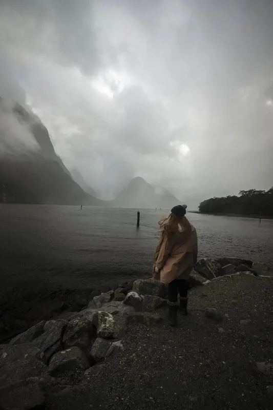 tasha amy standing on the rocky shore of Milford Sound on a cloudy and stormy day.